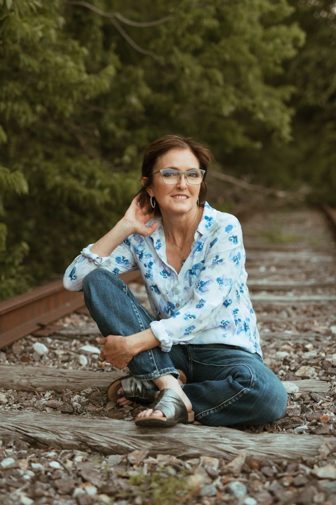 Subject sits casually on railroad tracks wearing blue floral top and jeans in autumn setting with fallen leaves.
