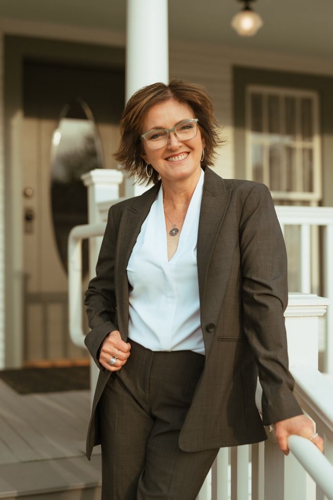 Professional in a brown business suit and glasses stands confidently on a front porch with white railing.