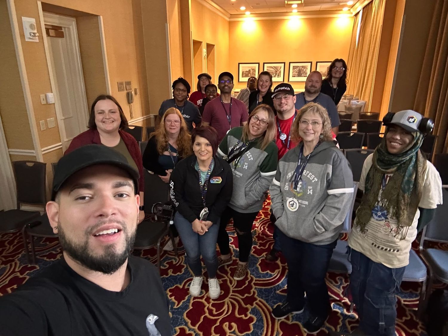 A group poses together for a selfie in a hotel hallway with patterned carpet and framed artwork on the walls.