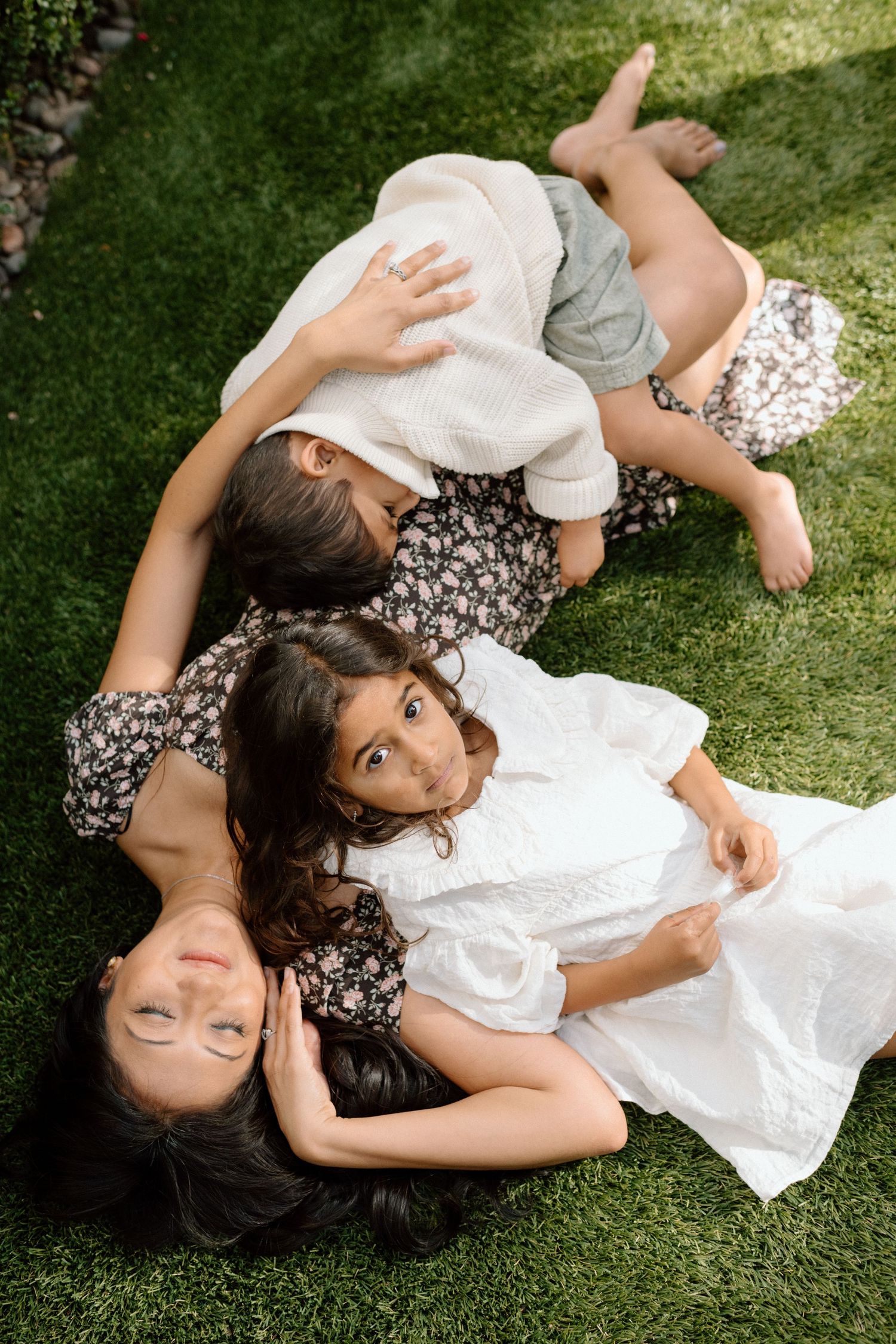 Three people in white and floral outfits laying on green grass in a natural outdoor setting.