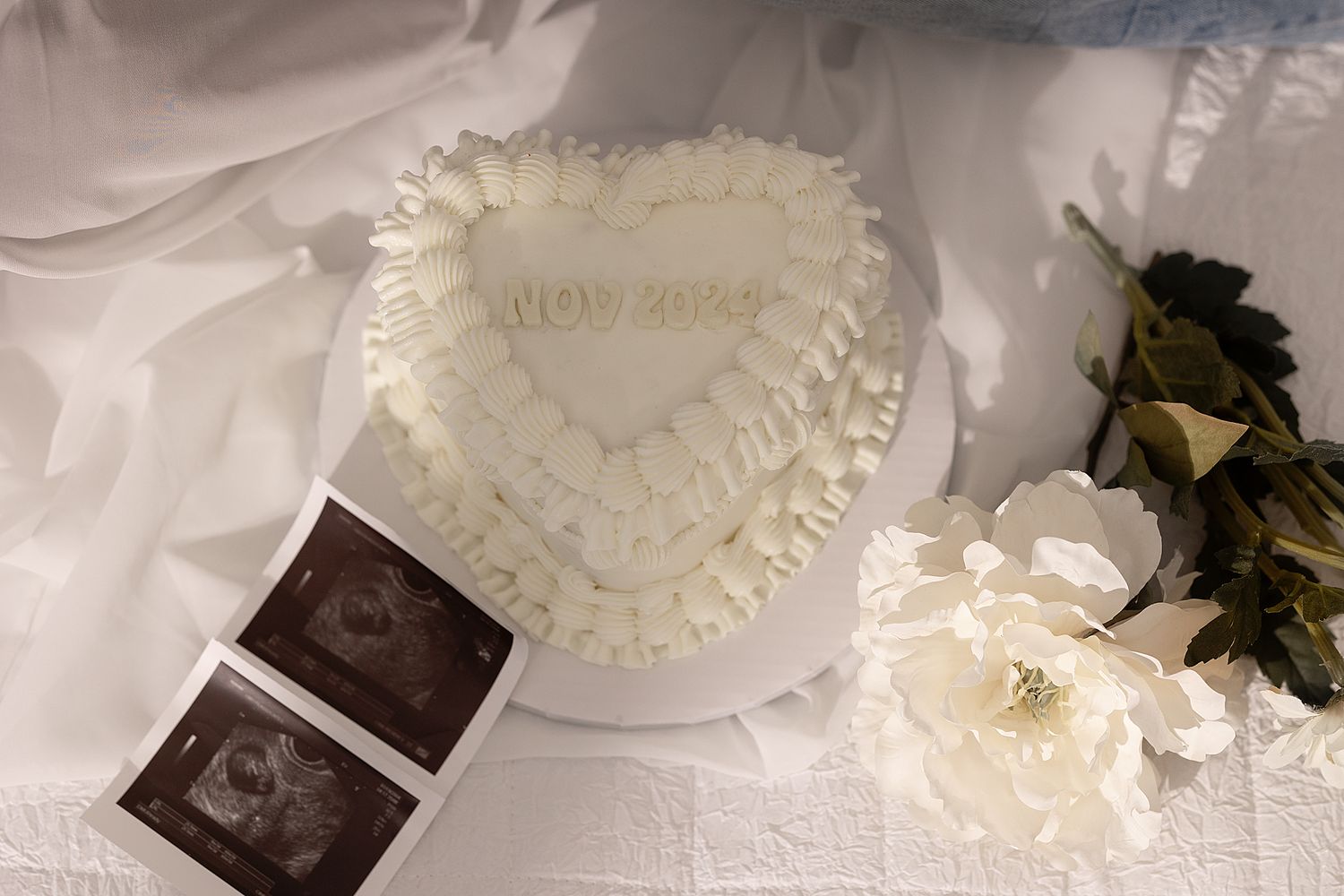 Gender Reveal-white heart-shaped cake next to ultrasound photos and white flowers on a light fabric background.