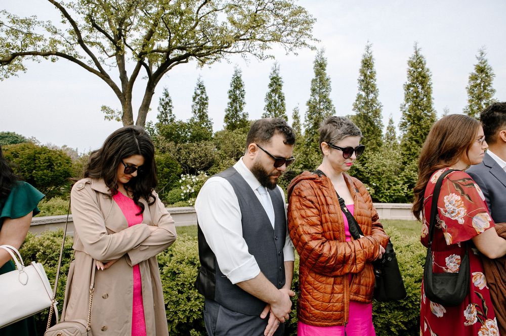 Group of party guests stand together outdoors in a garden setting looking at their phones.
