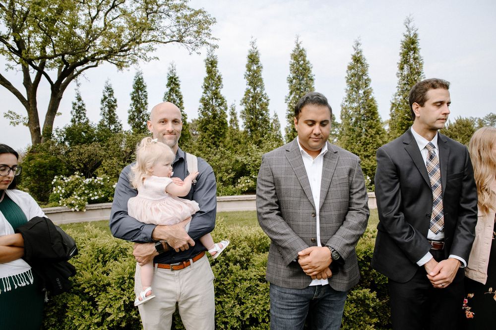 Wedding guests gather outside in a garden setting with tall evergreen trees and landscaping in the background.
