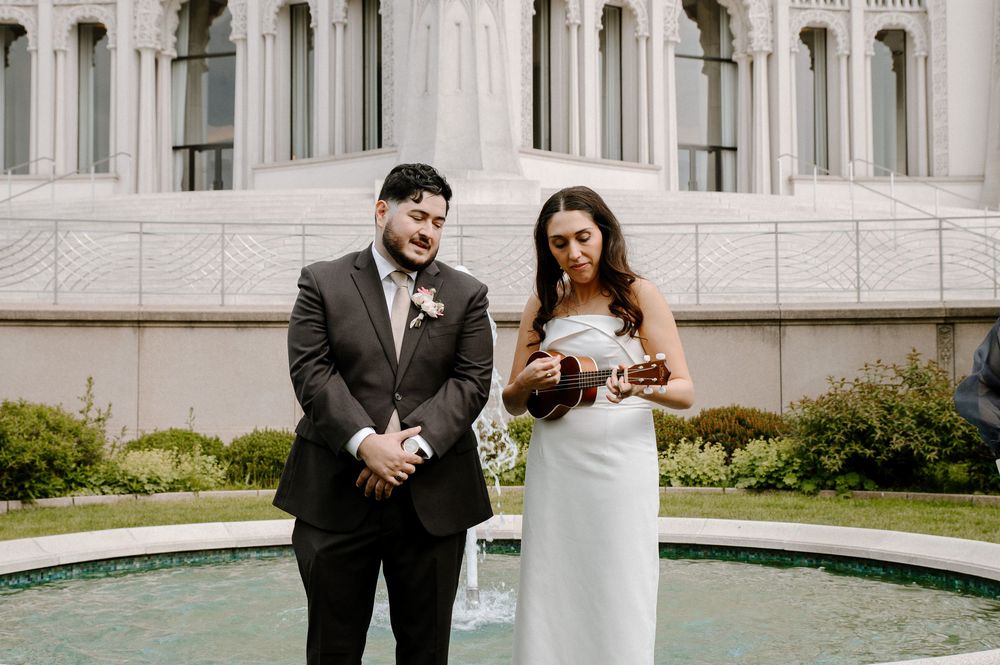 A couple stands together in formal wedding attire outside a grand white stone building with columns and fountains.