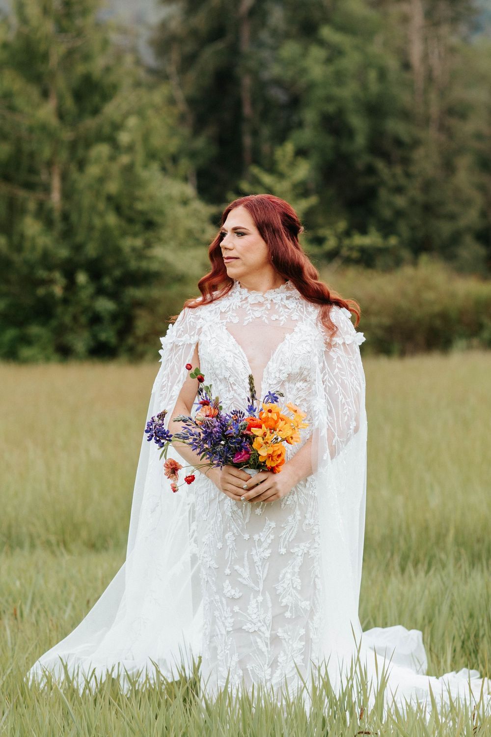 A bride in a white lace dress with long sleeves holds a vibrant purple and orange wildflower bouquet in a grassy meadow.
