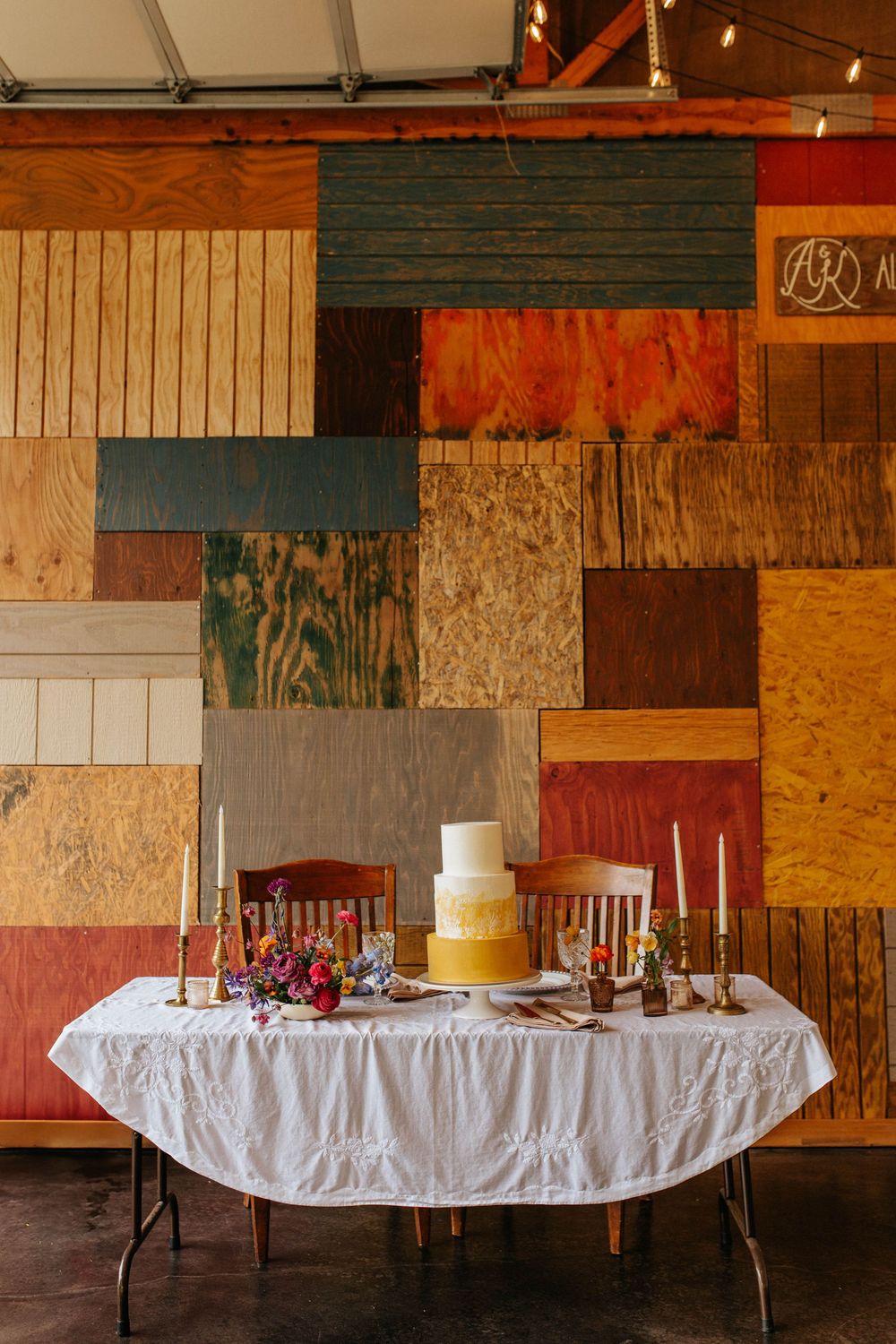 Rustic banquet tables with white tablecloths set up in sequence against a weathered wooden wall background.