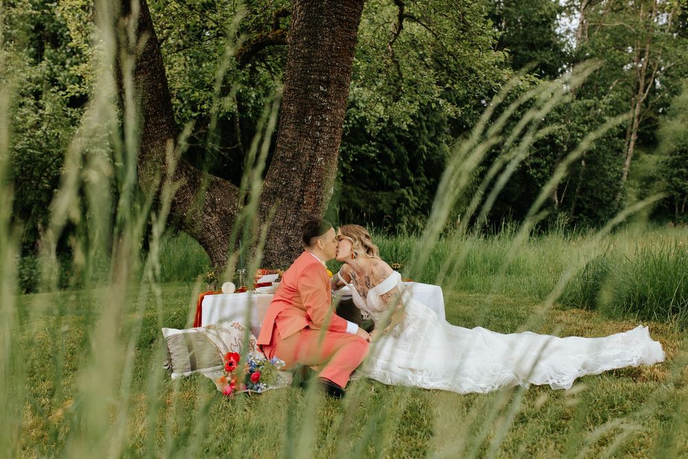 Two people recline on a white blanket for a romantic picnic surrounded by tall grass in a serene outdoor setting.