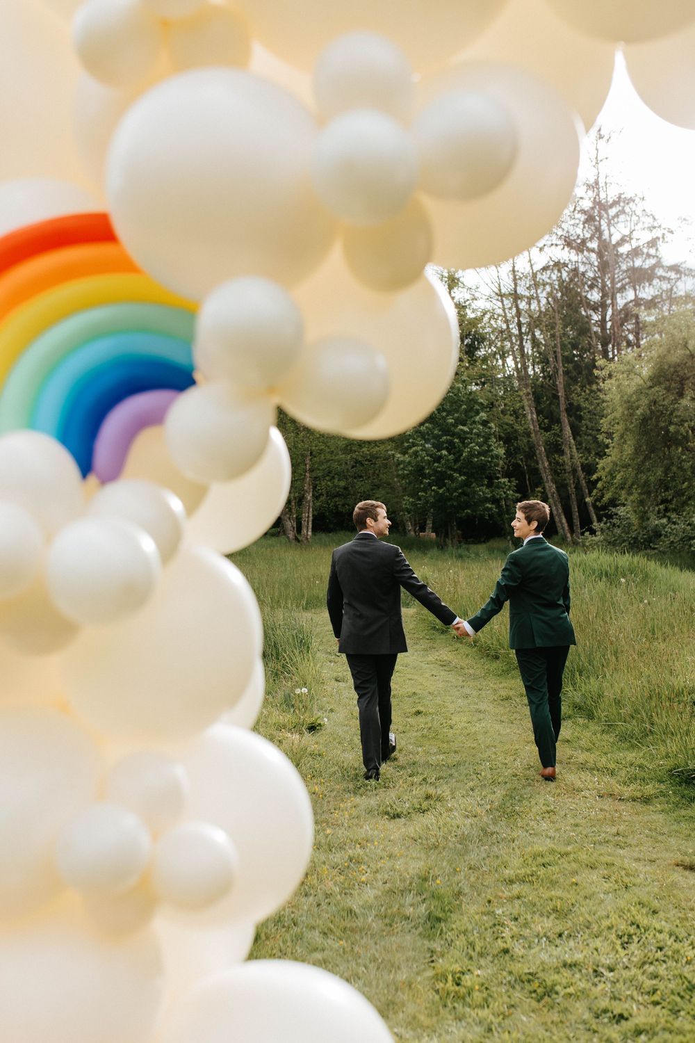 Two people in suits walk hand-in-hand on a grass path under a decorative rainbow and white balloon arch in nature.