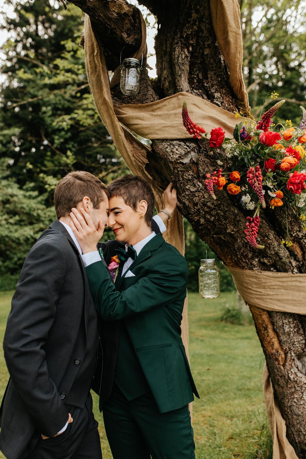 A romantic outdoor scene between two people wearing black and green formal attire beneath a rustic tree with floral decor.