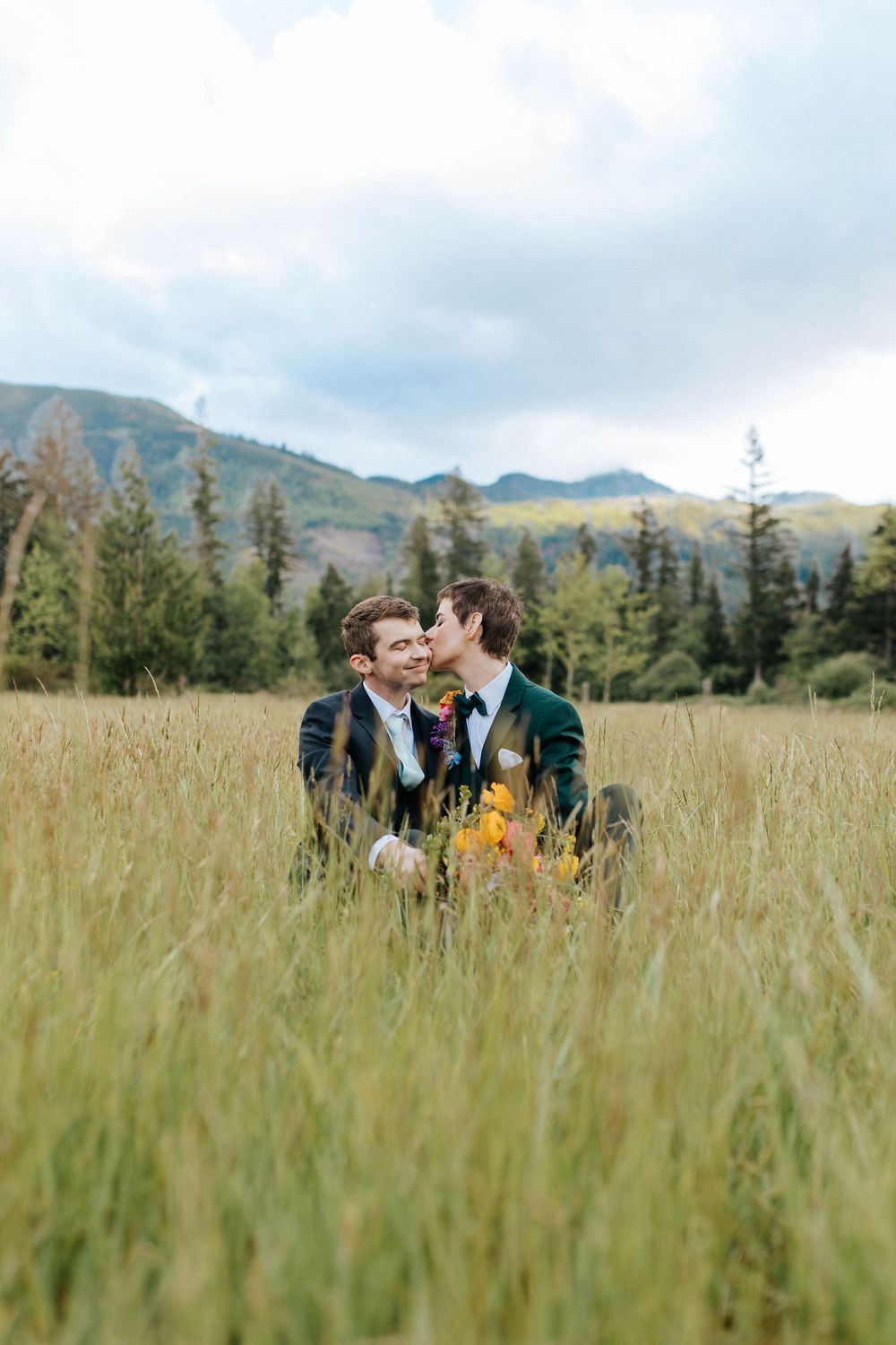Two people in suits share an intimate moment in a mountain meadow surrounded by evergreen trees.