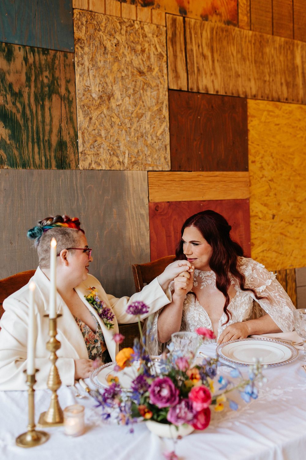 Two people share an intimate moment at a rustic wedding reception with colorful floral centerpiece and wooden wall backdrop.