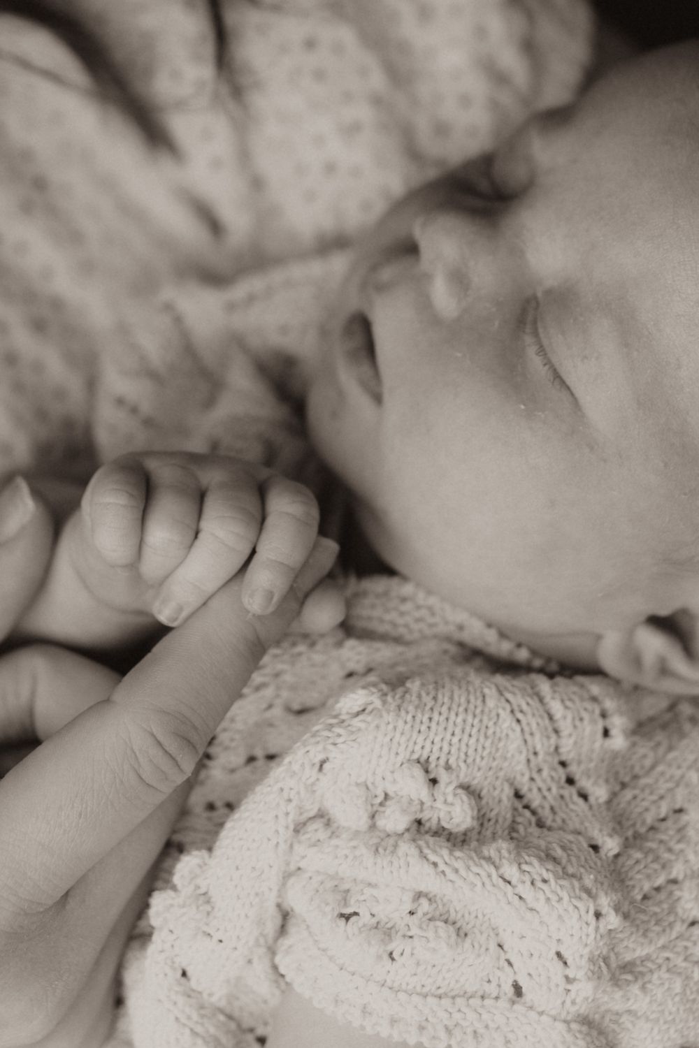 A sleeping baby in a white knitted sweater holds an adult finger in a tender black and white close-up photograph.