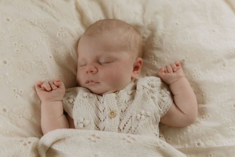 Peacefully sleeping infant wearing delicate white lace dress rests on cream colored textured bedding.