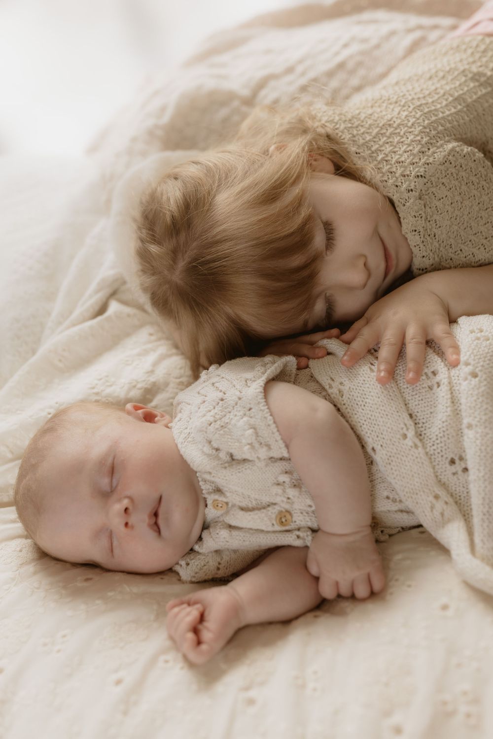 Two young children sleeping peacefully together on light colored bedding in a soft warm tone.