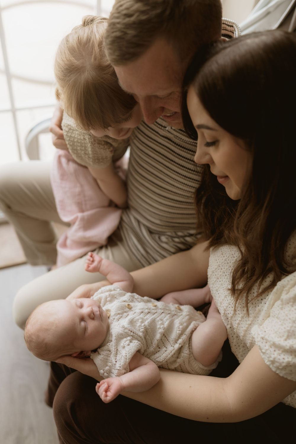 Family sits closely together on couch while holding and gazing lovingly at their newborn baby in white lace dress.