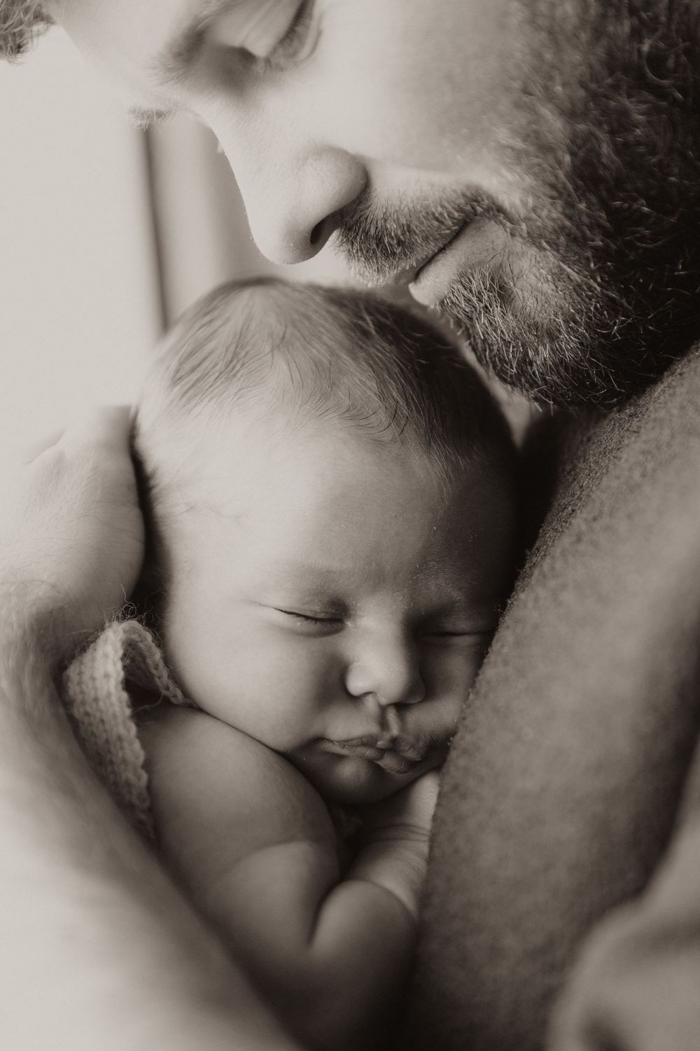 An intimate black and white portrait of a sleeping newborn baby being gently embraced by a bearded parent.