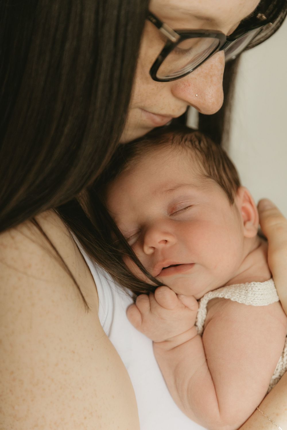 Intimate photo of peaceful sleeping infant nestled against caregiver wearing glasses in soft natural lighting.