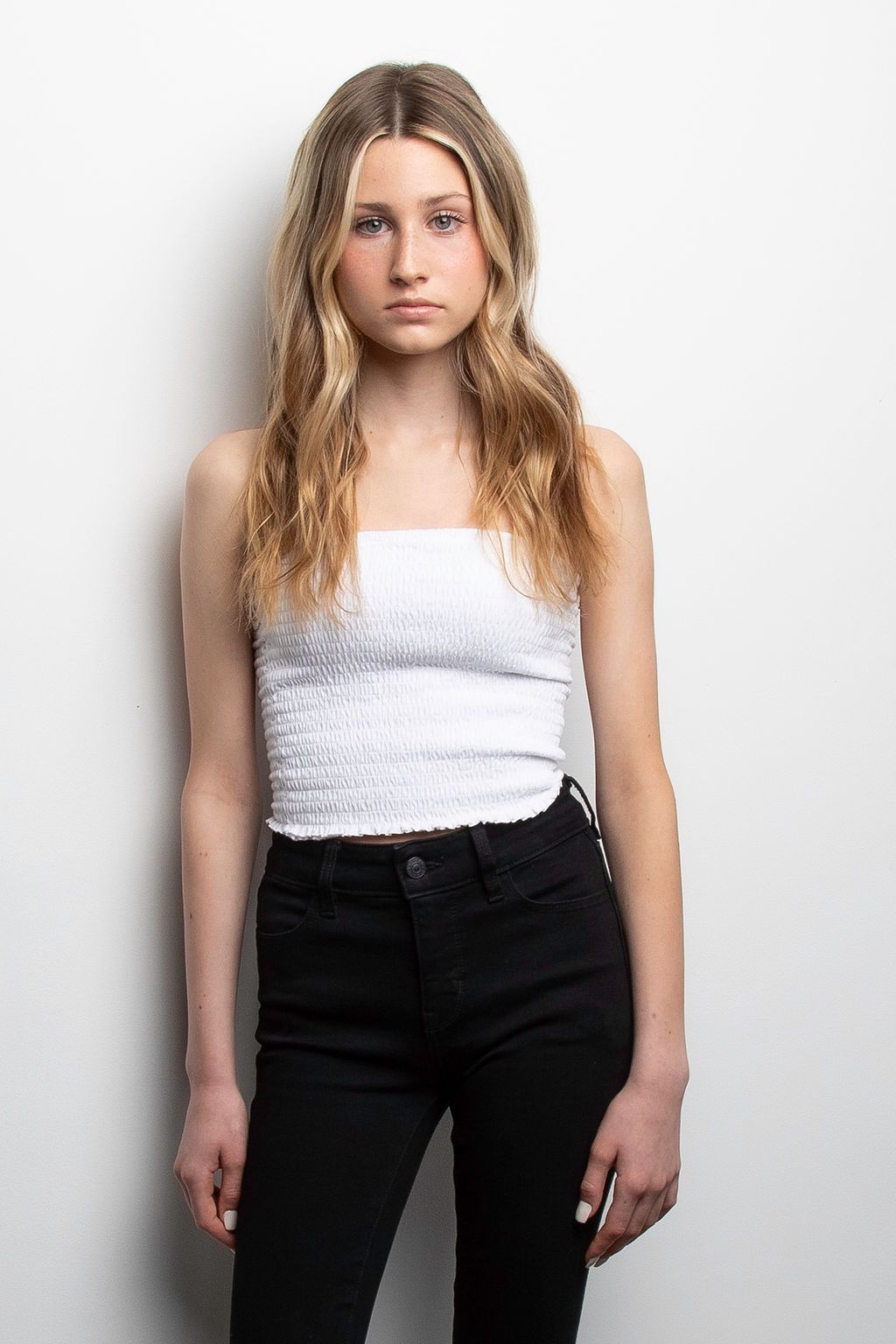 Fashion portrait with blonde wavy hair wearing white cropped top and black high-waisted jeans against light background.