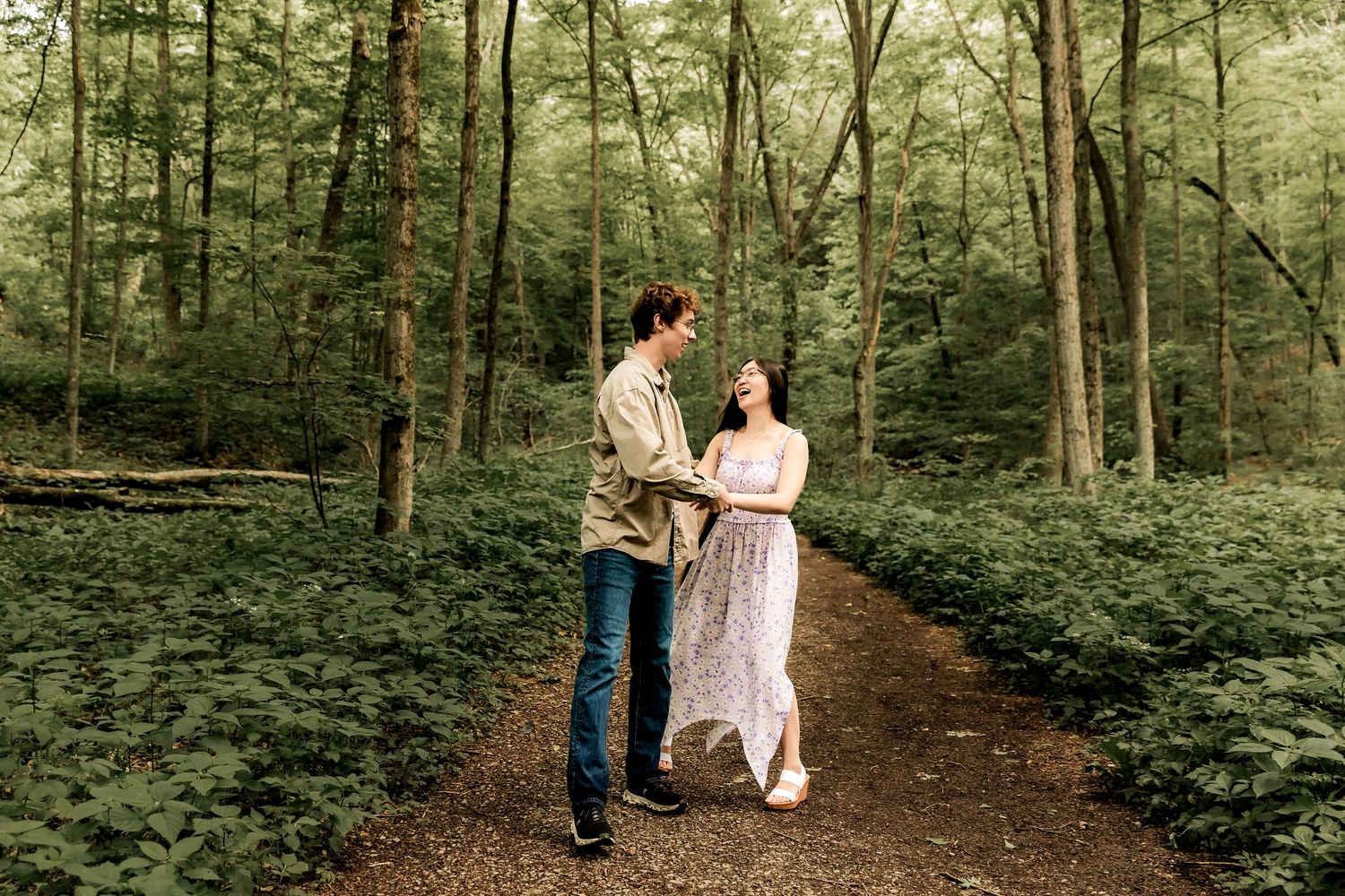 Two people hold hands while walking along a forest trail surrounded by lush green foliage.