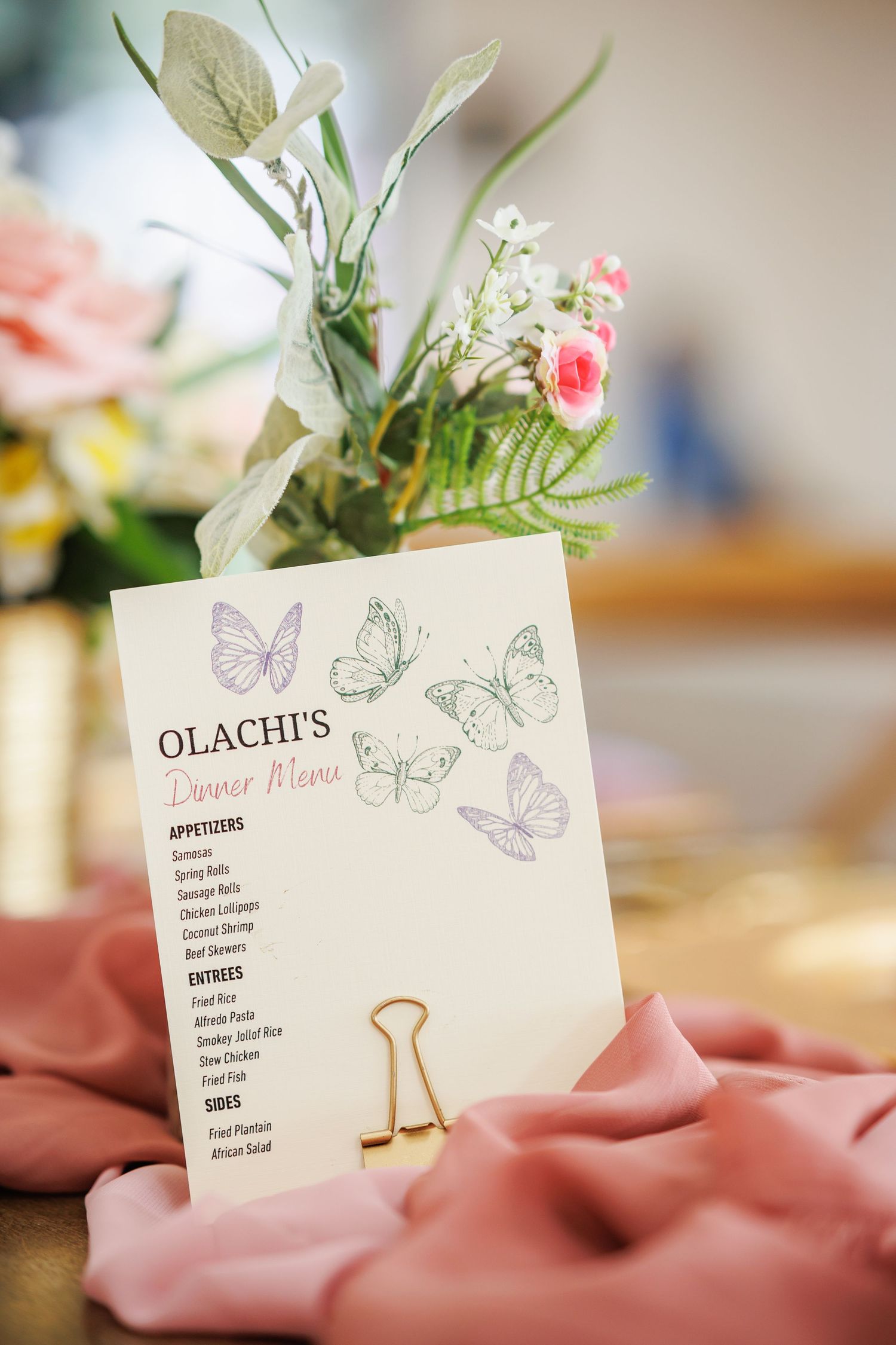 Birthday party dinner menu with butterfly designs held by a gold clip beside pink flowers and greenery on a table.