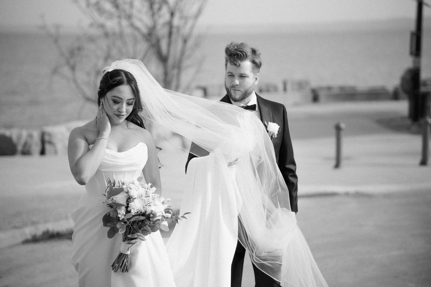 Wind catches bride's veil as couple shares candid moment outdoors in black and white.