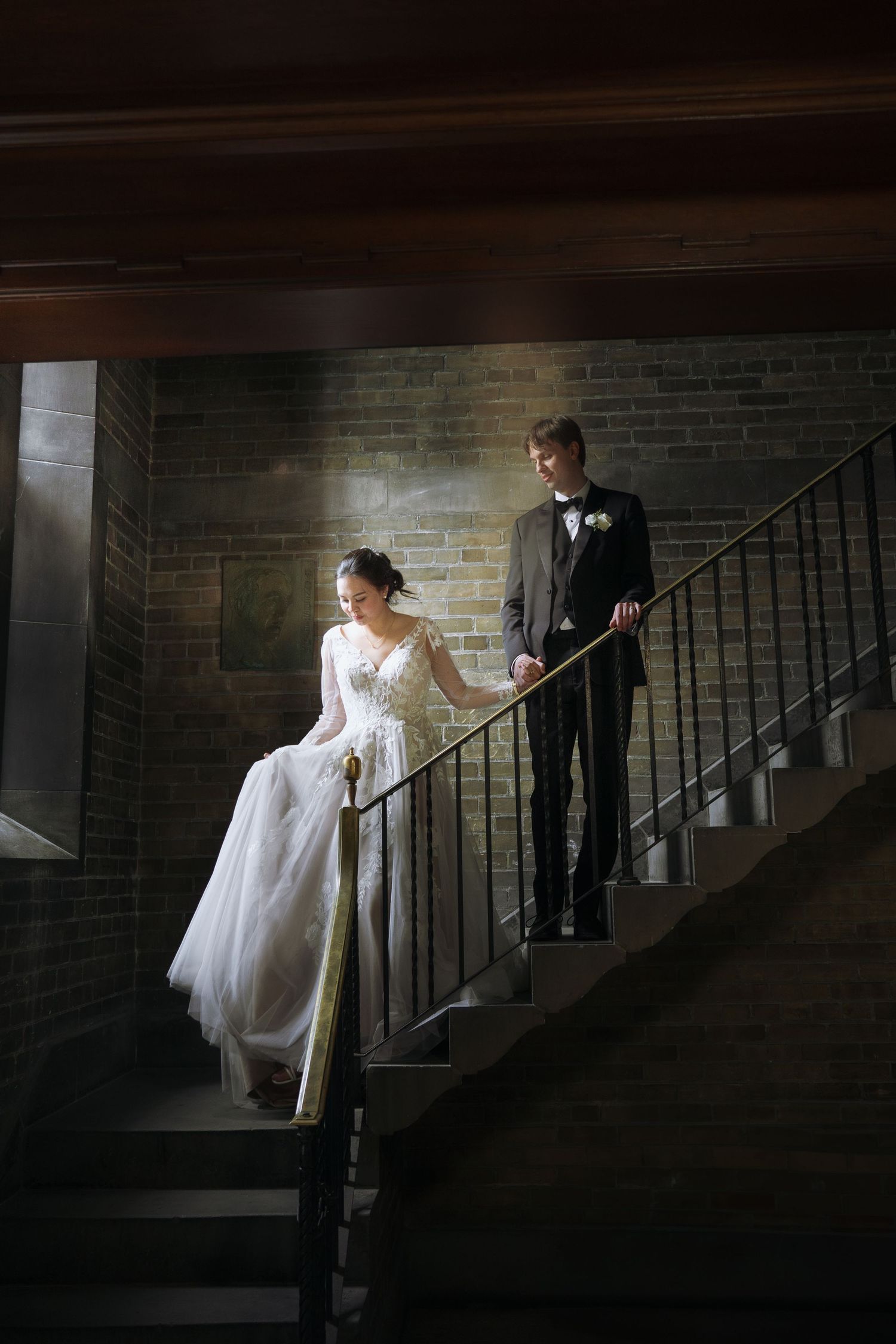 Dramatic moody staircase scene showcasing a white wedding dress and formal attire against brick walls.