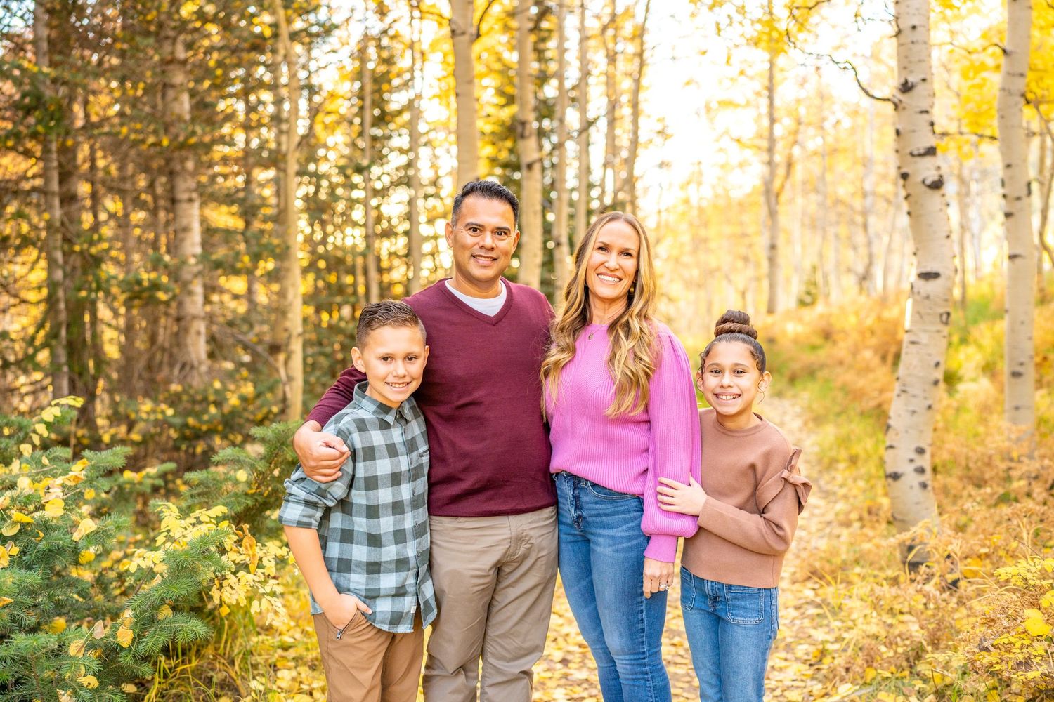 A group of four people pose together in a sunlit autumn forest with golden aspen trees and warm fall colors.