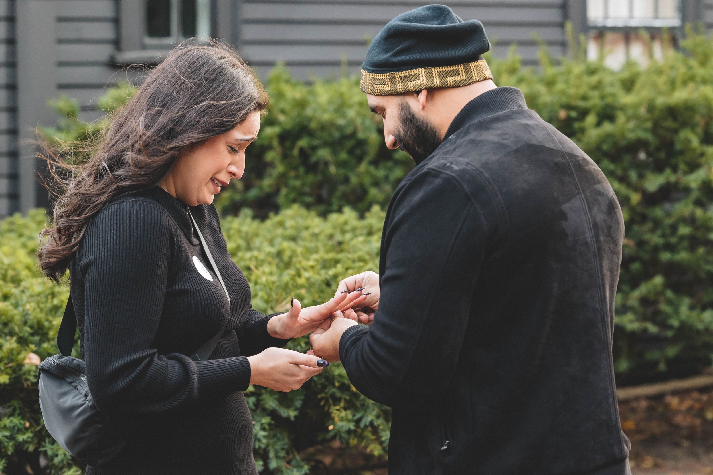A couple looks down at something being exchanged between them in front of shrubs.