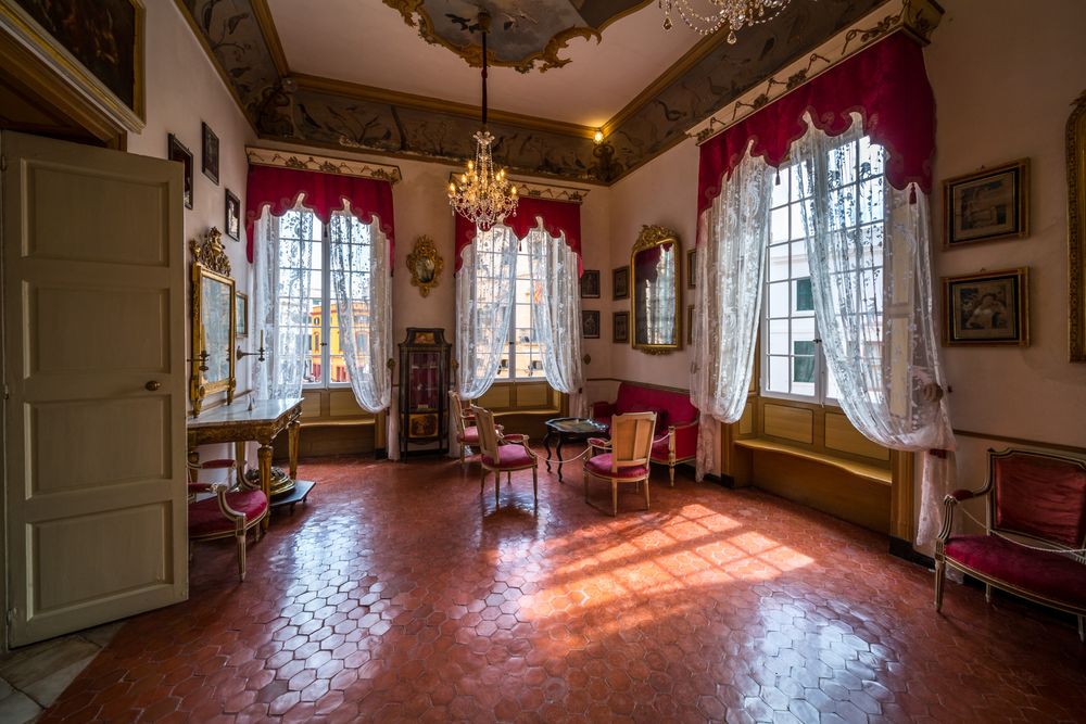 Sunlit Victorian parlor room with ornate windows, red curtains, antique furniture and terra cotta tile flooring.
