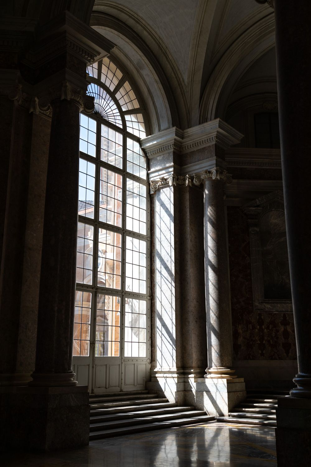 Sunlight streams through a tall arched window in an ornate palace interior with classical columns and marble floors.
