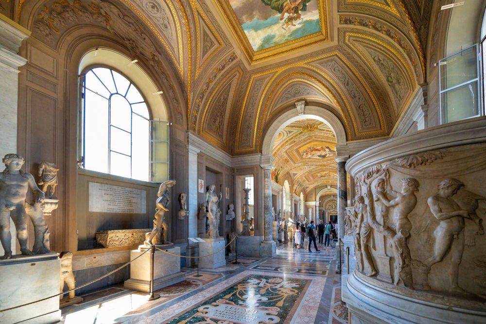 Ornate gallery corridor in Vatican Museums showcases classical sculptures and frescoed ceilings with marble floors and arches.