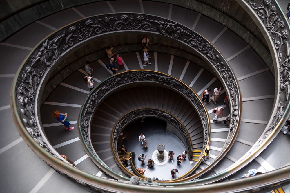 Spiral staircase in Vatican Museums with ornate railing and visitors seen from above walking down the steps.