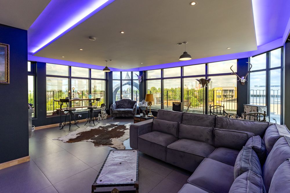 Modern luxury high-rise apartment living room with purple LED lighting, grey sectional sofa and panoramic city views.