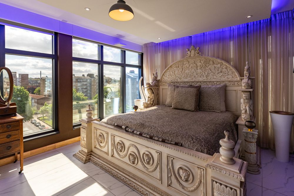 Modern bedroom with ornate white carved bed frame, gray bedding, floor-to-ceiling windows and blue ambient lighting.