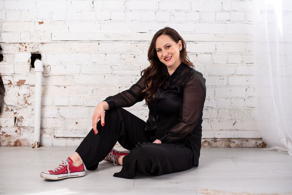 Seated figure in all-black outfit with sheer sleeves and red sneakers poses against white brick wall.