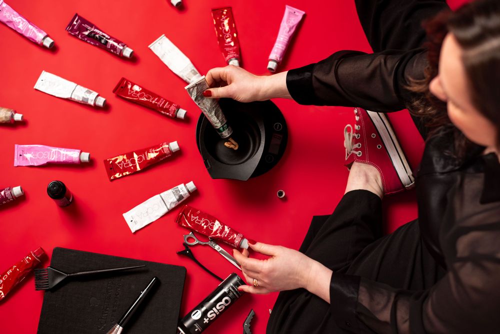 Beauty products and makeup tubes arranged in a circular pattern on a bright red surface with hands organizing items.