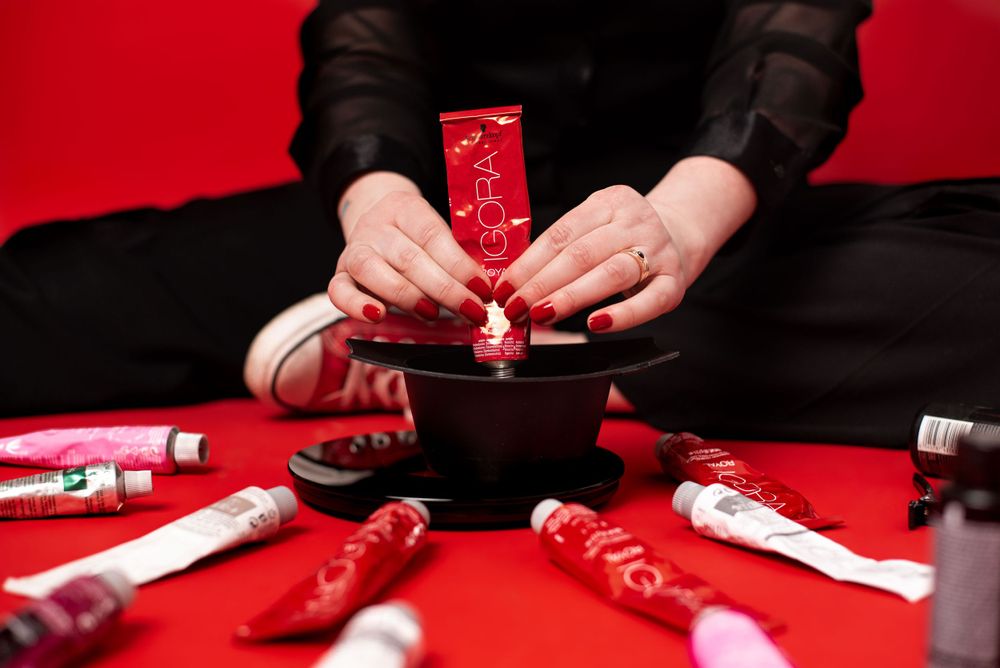 Hands arranging red beauty products and tubes around a black bowl against a vibrant red background.