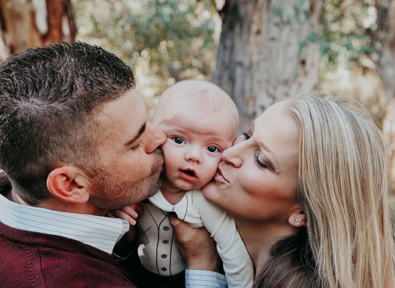 A beautiful outdoor family portrait of parents lovingly kissing their baby's cheeks during an autumn photoshoot.