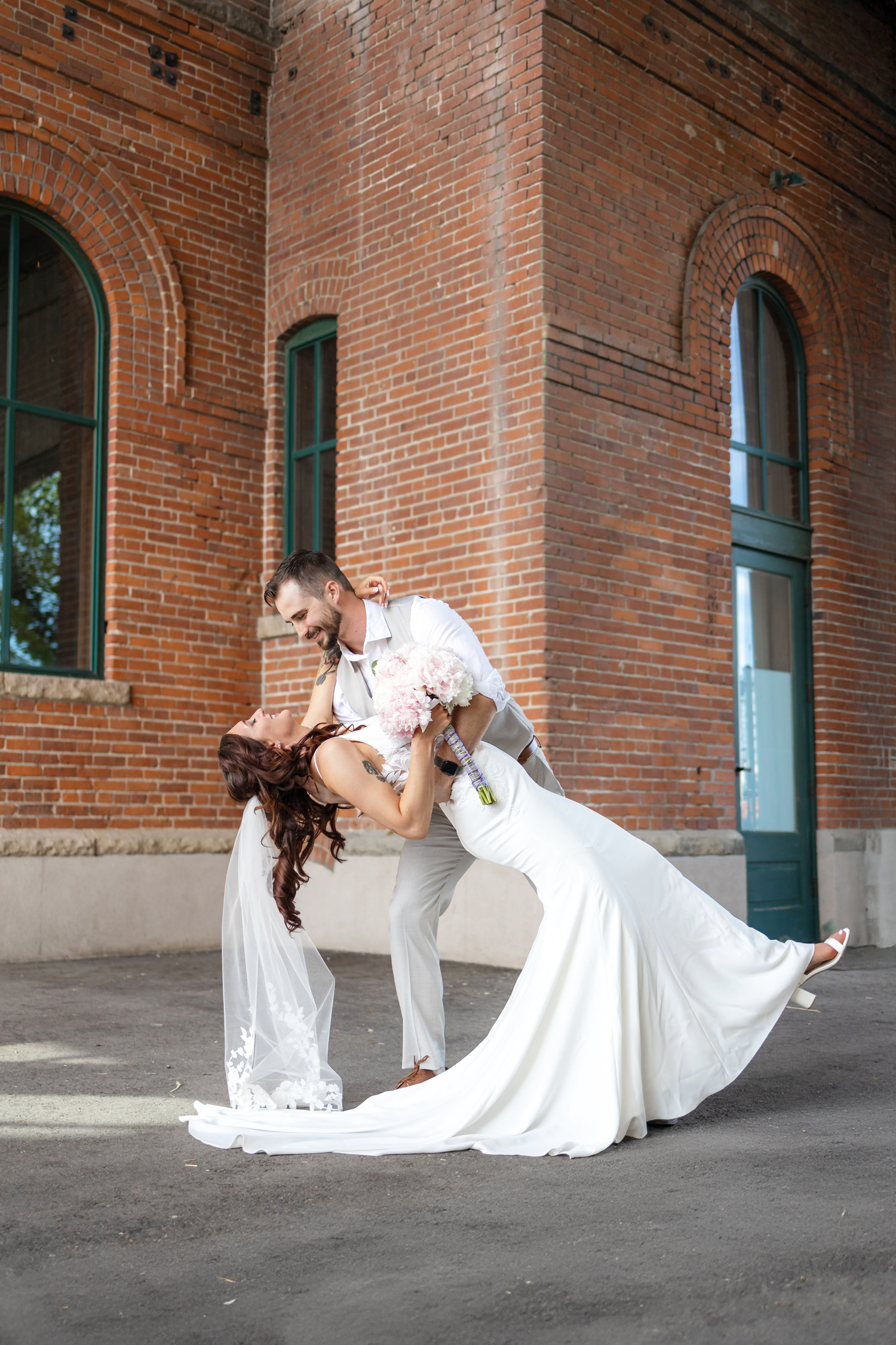 Romantic dip pose of couple in wedding attire against brick building exterior.