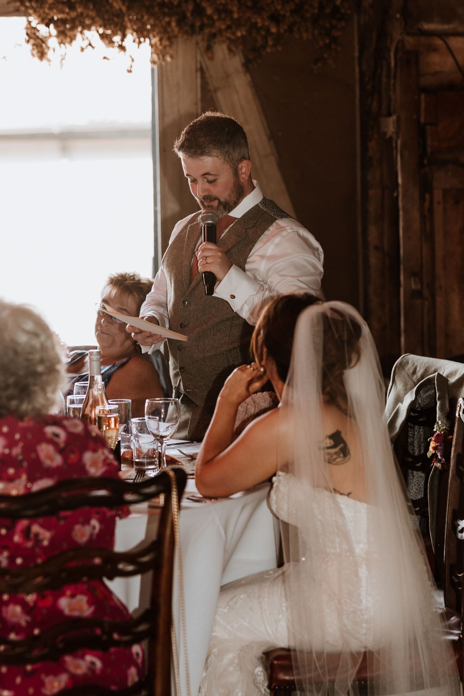 At a rustic wedding reception, a person in a vest gives an emotional speech while others listen at the dinner table at Lyde Court.