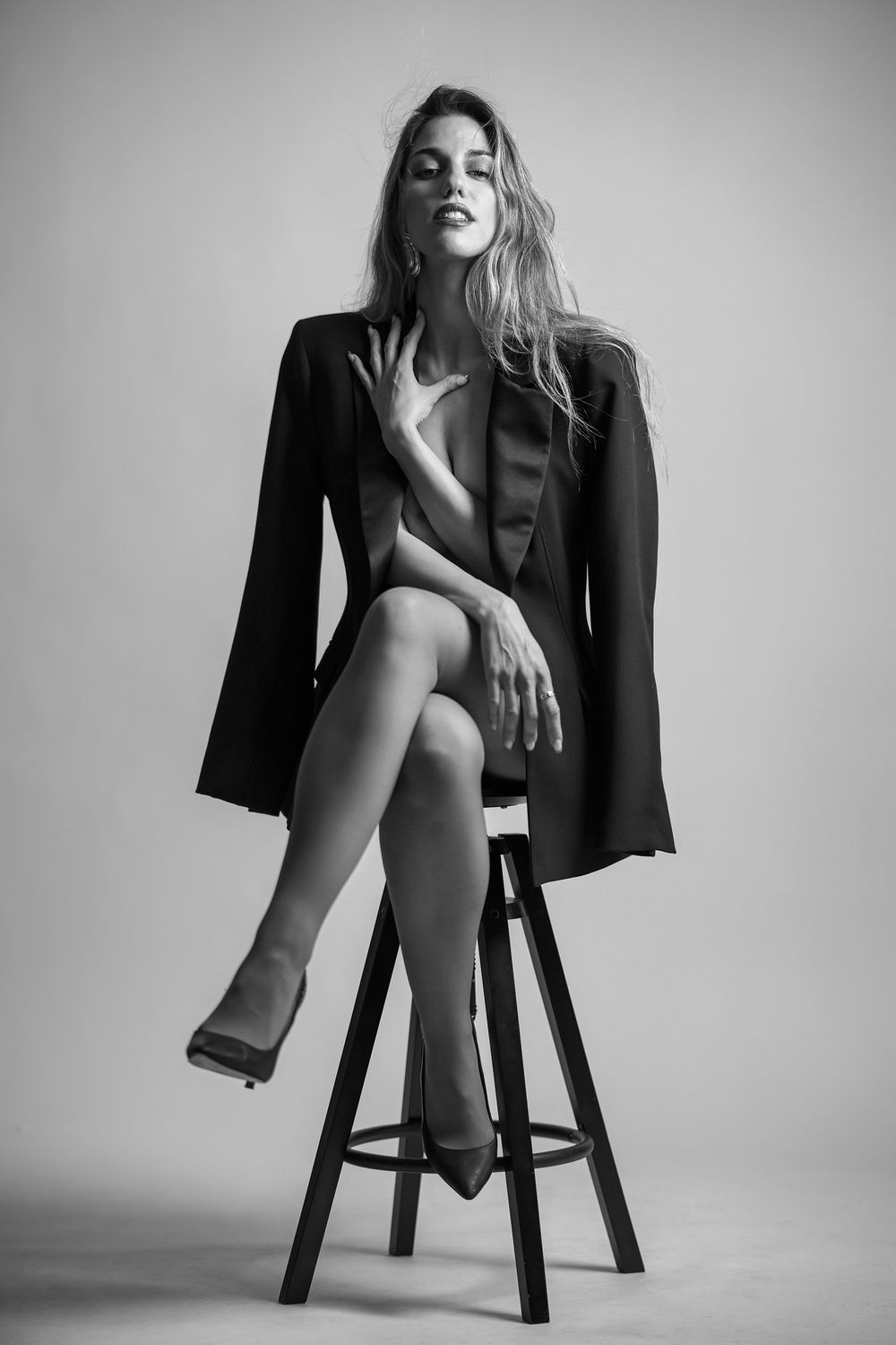 Black and white studio portrait of someone seated on a stool wearing an oversized blazer in an artistic pose.