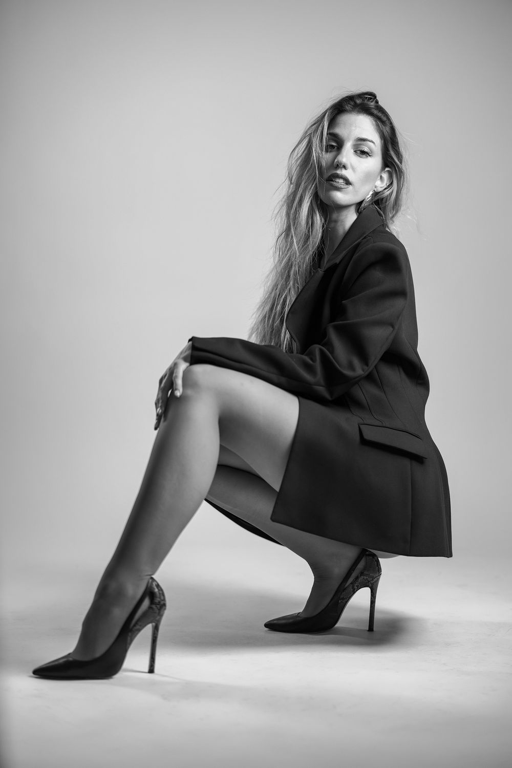 Black and white fashion portrait with person in blazer and high heels crouching in artistic pose against light backdrop.