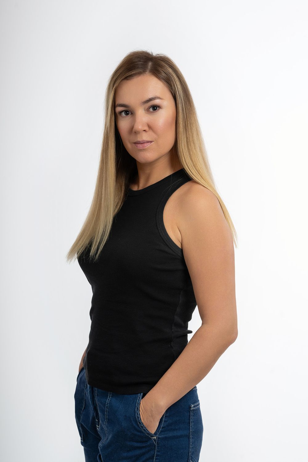 Studio portrait of a person with long blonde hair wearing a black sleeveless top and blue jeans against a white background.
