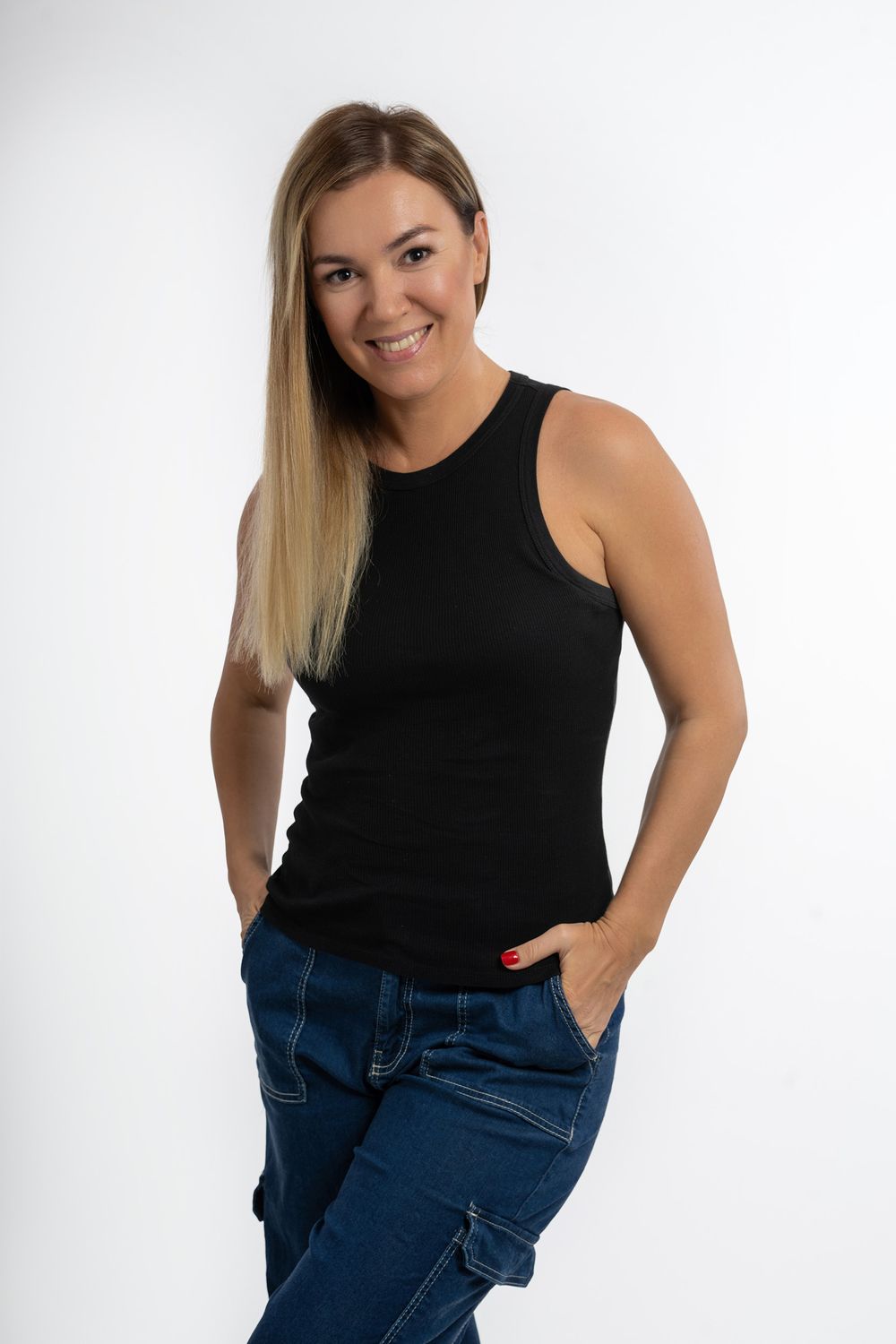 A woman in a black sleeveless top and blue jeans poses casually against a white background.