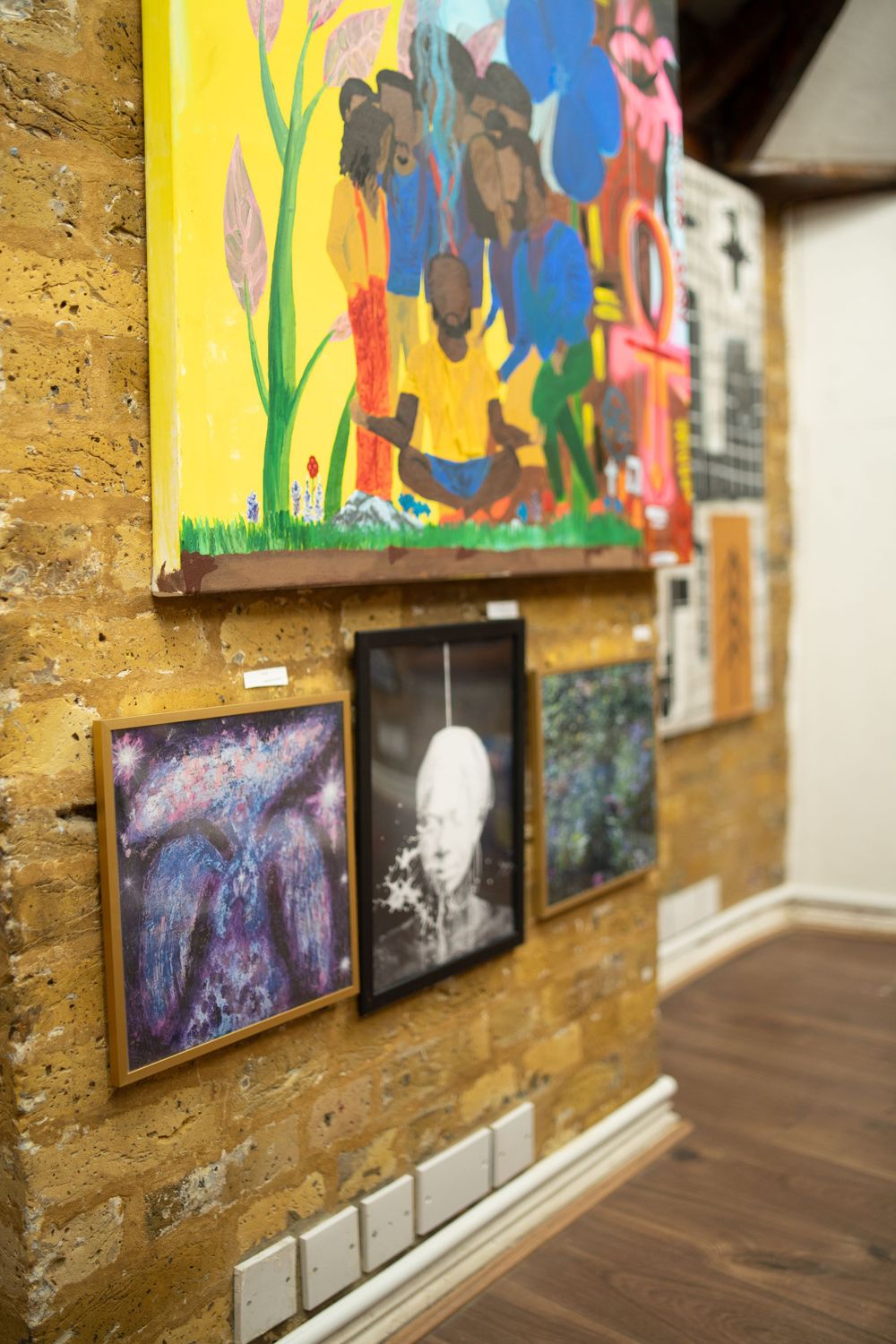 Framed artwork displayed on a yellow textured wall showing colorful paintings and photographs in a gallery setting.