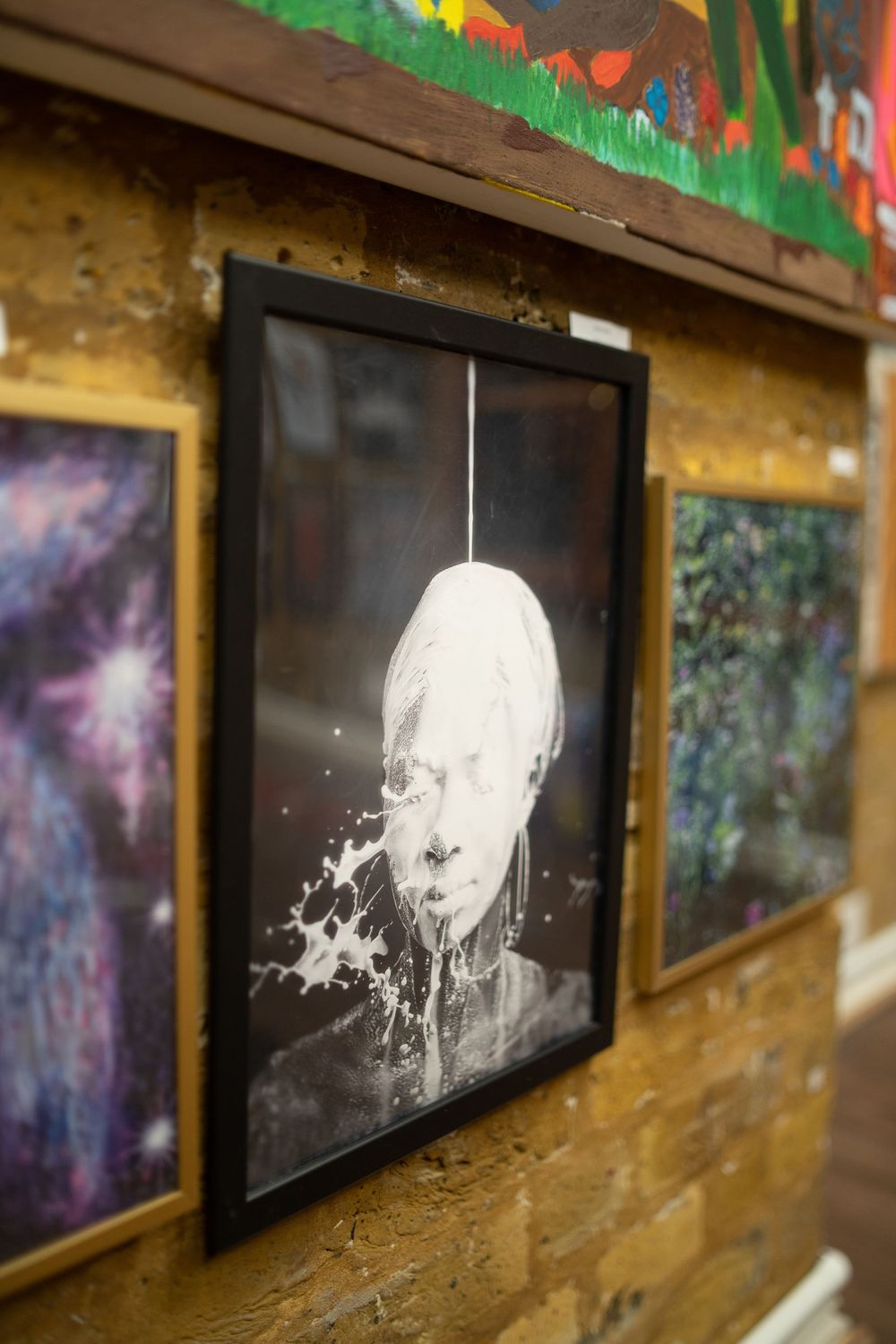 Black and white framed artwork showing a portrait with a milk splash effect mounted on textured yellow wall.