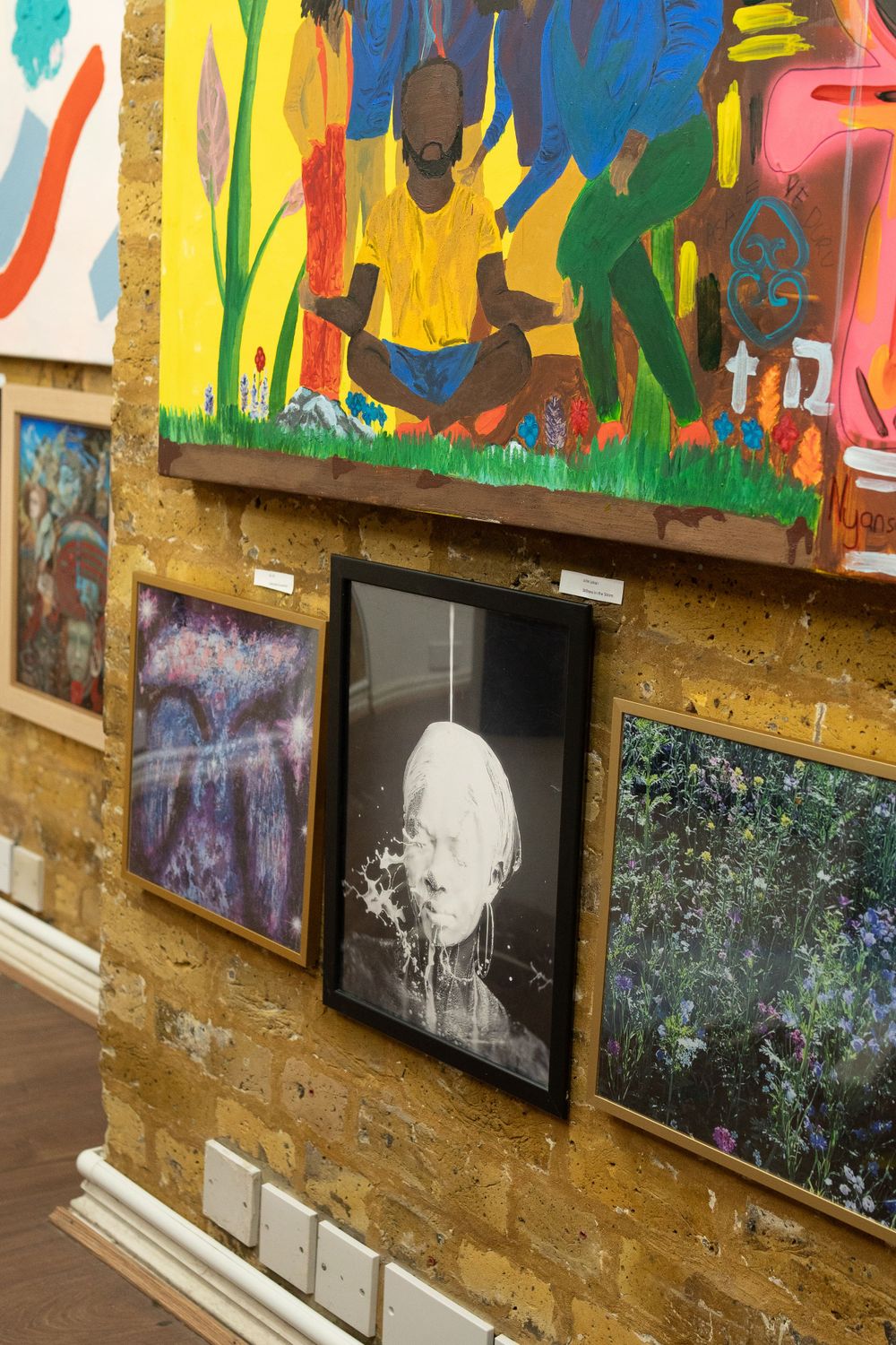 Art gallery wall displays colorful paintings and photographs mounted on a yellow textured surface.