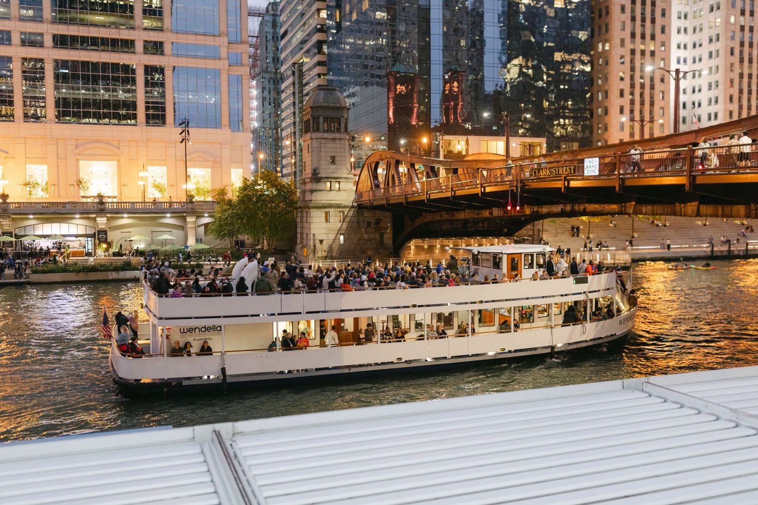 A river tour boat cruises along the Chicago River at dusk with illuminated skyscrapers and bridges in the background.