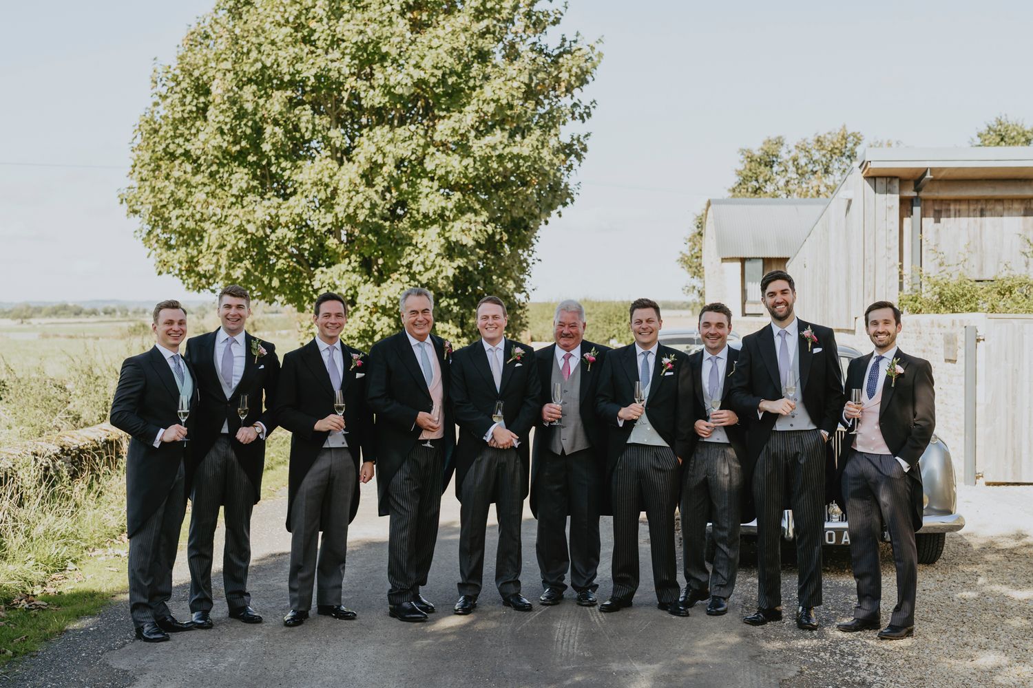 A group of groomsmen in black tuxedos and suits stand outdoors for a formal wedding photo.