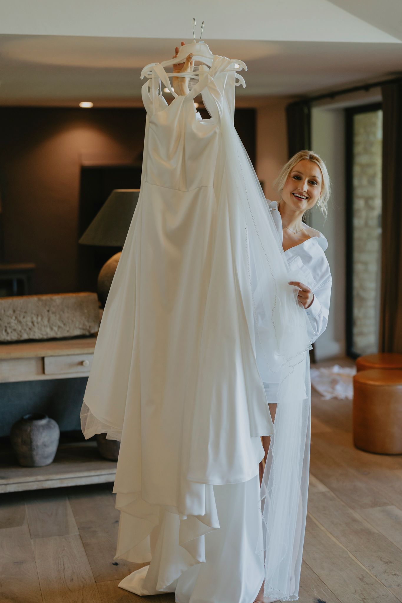 A flowing white wedding gown hangs elegantly while being held up in a sunlit room with wood floors.